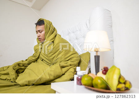 Indonesian southeast asian man in a blanket sitting on the bed at home and coughing. Suffer from fever, flu, and cold. Healthcare concept 127862313