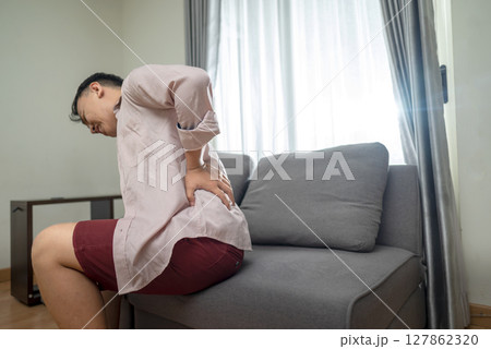 Indonesian southeast asian man suffering from back pain while sitting on the living room couch at home. Healthcare concept 127862320