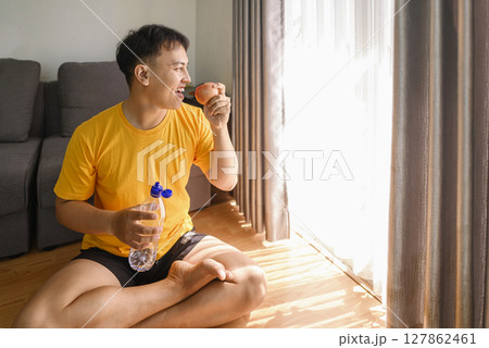 Indonesian southeast asian man drinking water and eating fruit after finishing a home body workout exercise session for a fit physique and healthy sport at home. Fitness and active lifestyles concept Indonesian southeast asian man drinking water and eating fruit after finishing a home body workout exercise session for a fit physique and healthy sport at home. Fitness and active lifestyles concept 127862461