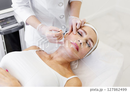 Mature woman getting rejuvenation injection in beauty clinic by cosmetologist in protective gloves and white coat, anti-aging facial treatment to smooth wrinkles and restore youthful skin appearance 127862499