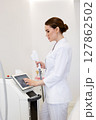 Woman cosmetologist in white coat adjusting laser device before non-surgical skin rejuvenation procedure in beauty clinic, modern equipment for aesthetic dermatology and anti-aging facial treatment 127862502
