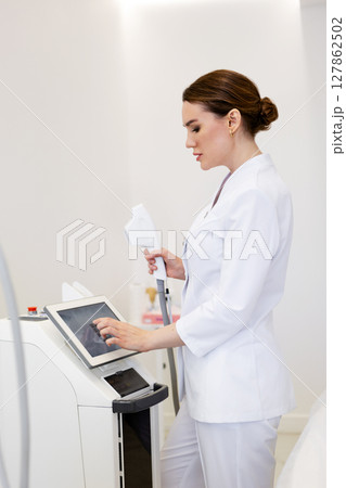 Woman cosmetologist in white coat adjusting laser device before non-surgical skin rejuvenation procedure in beauty clinic, modern equipment for aesthetic dermatology and anti-aging facial treatment Woman cosmetologist in white coat adjusting laser device before non-surgical skin rejuvenation procedure in beauty clinic, modern equipment for aesthetic dermatology and anti-aging facial treatment 127862502