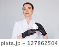 Woman cosmetologist in white coat holding blank product box in black glove, promoting professional skincare or beauty treatment in modern clinic, isolated on light background 127862504