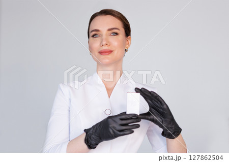 Woman cosmetologist in white coat holding blank product box in black glove, promoting professional skincare or beauty treatment in modern clinic, isolated on light background 127862504