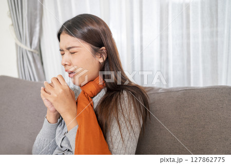 Indonesian southeast asian woman in a scarf sitting on the couch at home and coughing. Suffer from fever, flu, and cold. Healthcare concept Indonesian southeast asian woman in a scarf sitting on the couch at home and coughing. Suffer from fever, flu, and cold. Healthcare concept 127862775