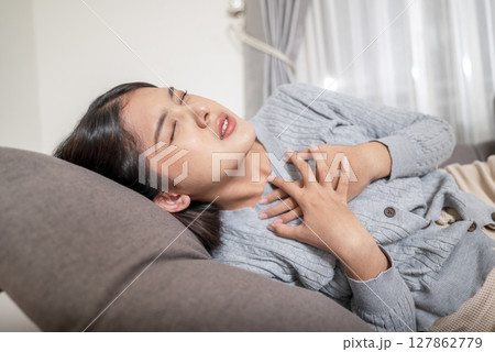 Indonesian southeast asian woman lying down on the couch and suffering from chest pain and touching her heart area. Pressure in the chest. Healthcare concept Indonesian southeast asian woman lying down on the couch and suffering from chest pain and touching her heart area. Pressure in the chest. Healthcare concept 127862779