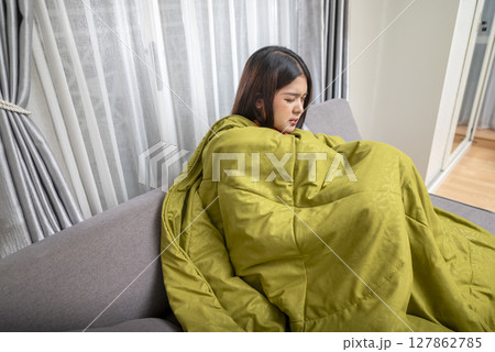 Indonesian southeast asian woman in a blanket sitting on the couch at home and coughing. Suffer from fever, flu, and cold. Healthcare concept Indonesian southeast asian woman in a blanket sitting on the couch at home and coughing. Suffer from fever, flu, and cold. Healthcare concept 127862785