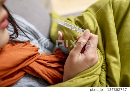 Indonesian southeast asian woman in a blanket holding a thermometer while sitting on the couch at home and coughing. Suffer from fever, flu, and cold. Healthcare concept Indonesian southeast asian woman in a blanket holding a thermometer while sitting on the couch at home and coughing. Suffer from fever, flu, and cold. Healthcare concept 127862787