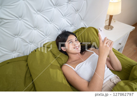 Indonesian southeast asian woman lying down on a bed and using a cellphone in the morning after waking up at home 127862797
