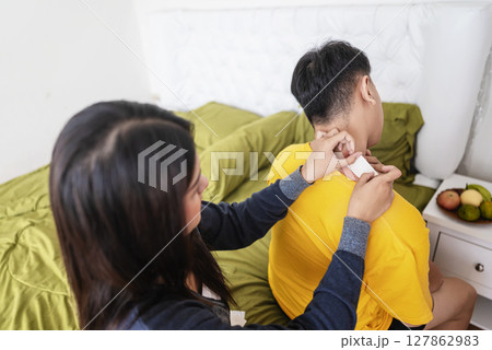 Indonesian southeast asian woman applies a neck pain patch to her husband neck in the bedroom. Self-care and wellness routines concept Indonesian southeast asian woman applies a neck pain patch to her husband neck in the bedroom. Self-care and wellness routines concept 127862983