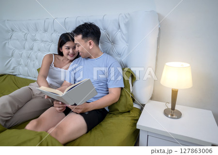 Indonesian southeast asian couple lying on a bed together while reading a book in the bedroom at night before sleep 127863006