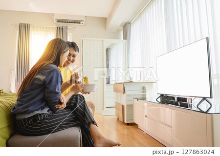 Indonesian southeast asian couple family eating noodles meal together while watching television at home. Empty television screen for mockup, template, or copy space Indonesian southeast asian couple family eating noodles meal together while watching television at home. Empty television screen for mockup, template, or copy space 127863024
