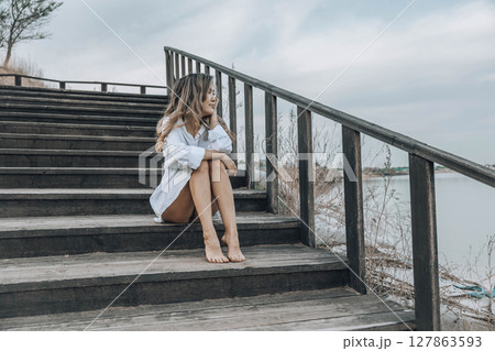Asian woman sitting barefoot on wooden staircase near lake, gazing into the distance with a relaxed and confident expression, wearing white shirt dress, travel, lifestyle, selfcare 127863593