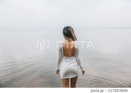 unrecognizable woman standing with back exposed in open white dress near calm lake, facing the water with relaxed posture and serene mood, concept of wellness, spa, fashion editorial unrecognizable woman standing with back exposed in open white dress near calm lake, facing the water with relaxed posture and serene mood, concept of wellness, spa, fashion editorial 127863606