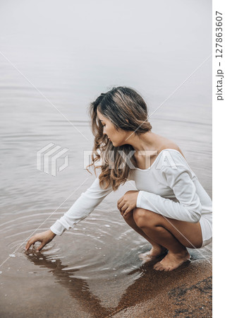 Asian woman squatting barefoot at lake edge creating water ripples with fingers, gazing at water with serene expression, calm horizon under cloudy sky, mindfulness, wellness, retreat Asian woman squatting barefoot at lake edge creating water ripples with fingers, gazing at water with serene expression, calm horizon under cloudy sky, mindfulness, wellness, retreat 127863607