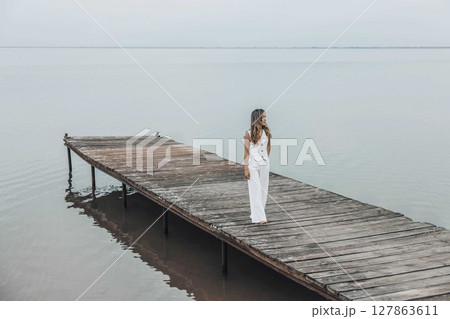 Asian woman in white outfit stands on wooden pier, calm lake, peaceful overcast, viewed from behind with contemplative mood, mindfulness, travel retreats, wellness Asian woman in white outfit stands on wooden pier, calm lake, peaceful overcast, viewed from behind with contemplative mood, mindfulness, travel retreats, wellness 127863611
