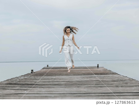 Asian woman jumping barefoot on wooden pier, calm sea horizon, dressed in white outfit under pale cloudy sky, mindfulness, wellness retreat, travel blog, motion blur Asian woman jumping barefoot on wooden pier, calm sea horizon, dressed in white outfit under pale cloudy sky, mindfulness, wellness retreat, travel blog, motion blur 127863613