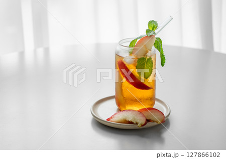 Ice tea with peach, mint in a glass on a white table near window with slice fruits and sunlight. Ice tea with peach, mint in a glass on a white table near window with slice fruits and sunlight. 127866102