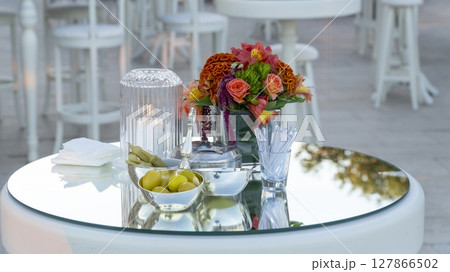 An elegant table setting with a floral centerpiece, olives, pickles, and drink stirrers on a reflective surface, with blurred chairs and tables in background 127866502