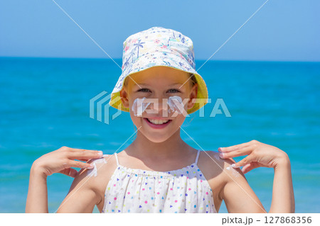 Child happily applying sunscreen on her face and shoulders at the beach, wearing a vibrant hat Child happily applying sunscreen on her face and shoulders at the beach, wearing a vibrant hat 127868356