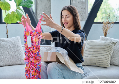 Young woman unpacking cardboard box parcel with online order, looking at new clothes 127869802