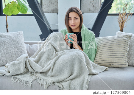 Beautiful young woman sitting under warm blanket with cup in her hands, on sofa 127869804