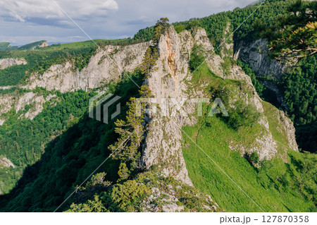 Dramatic rocky cliff surrounded by lush forest and green hills under a summer sky, captured in daylight. 127870358
