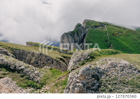 Mountain ridge with green slopes and dramatic cliffs rising above clouds on a moody summer day. Bolshoy Tkhach national park 127870360