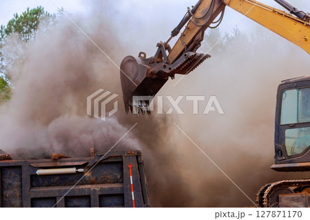 Heavy machinery operates to load construction debris into truck, creating cloud of dust at works site. 127871170