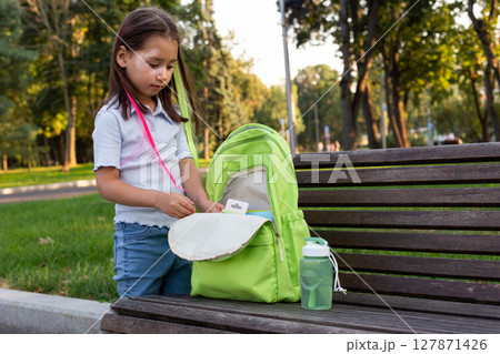 Young girl packing backpack in park with water bottle Young girl packing backpack in park with water bottle 127871426