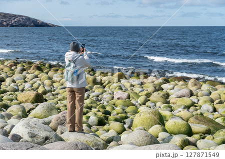 Woman in light hiking outfit takes a photo of the Barents Sea on Dragon Eggs Beach in Teriberka, standing among round moss-covered boulders 127871549
