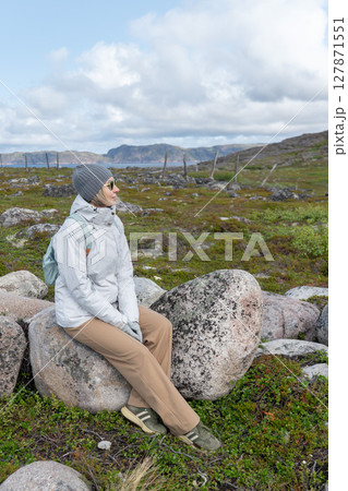 Smiling woman in a white jacket sitting on a large stone surrounded by tundra landscape, with mountains and cloudy sky in the background 127871551