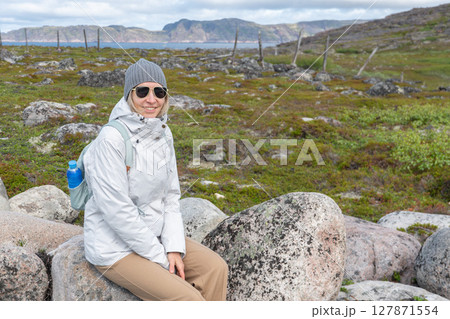 Smiling woman in a gray beanie, white jacket, and sunglasses sitting on a large granite rock in the Teriberka Nature Reserve tundra with scenic hills 127871554