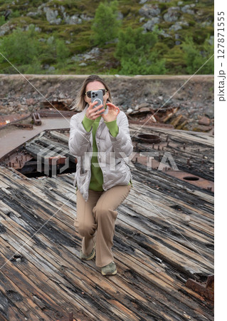 Woman taking a photo with a smartphone on the wooden wreck of an old ship surrounded by rugged northern landscape 127871555