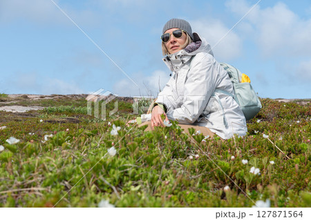 Woman in a gray beanie, white jacket, and sunglasses sitting among arctic vegetation in Teriberka Nature Reserve under a clear blue sky Woman in a gray beanie, white jacket, and sunglasses sitting among arctic vegetation in Teriberka Nature Reserve under a clear blue sky 127871564