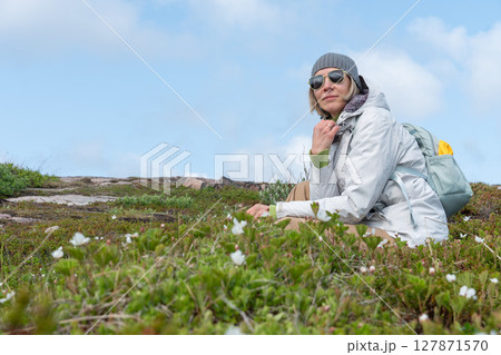 Woman in a gray beanie, white jacket, and sunglasses sitting among arctic vegetation in Teriberka Nature Reserve under a clear blue sky Woman in a gray beanie, white jacket, and sunglasses sitting among arctic vegetation in Teriberka Nature Reserve under a clear blue sky 127871570