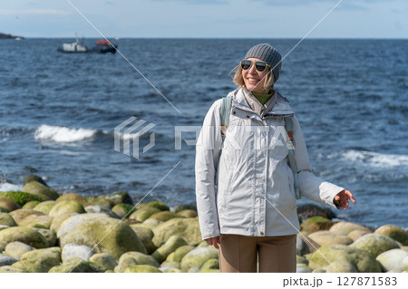 Woman smiles while standing on the distinctive rounded boulders at Dragon Eggs Beach in Teriberka, with the Barents Sea and a boat in the background 127871583