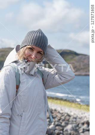 Woman in a gray knit hat and light jacket smiles as she holds her hair in the wind on a rocky Arctic beach 127871608