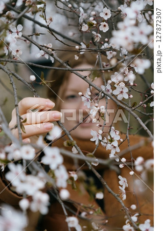 woman with red hair stands in front of a tree with pink flowers. She's wearing a brown coat, scarf. 127872307