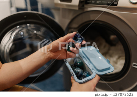 Close-up of unrecognizable man holding two laundry detergent pods preparing to load washing machine with clothes for fresh and clean laundry. Concept of cleanliness and housekeeping. Close-up of unrecognizable man holding two laundry detergent pods preparing to load washing machine with clothes for fresh and clean laundry. Concept of cleanliness and housekeeping. 127872522