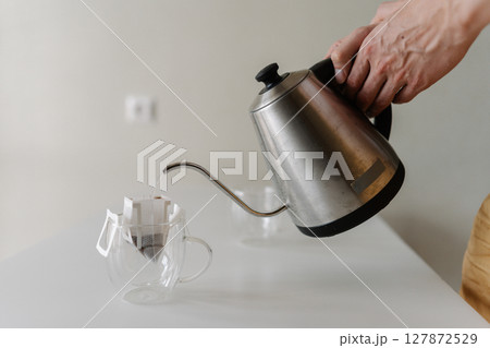 Close-up of unrecognizable young man preparing drip coffee bag by pouring hot water from stainless steel kettle into clear glass cup placing on white table at home, creating inviting morning ritual. Close-up of unrecognizable young man preparing drip coffee bag by pouring hot water from stainless steel kettle into clear glass cup placing on white table at home, creating inviting morning ritual. 127872529