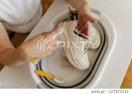 Close-up cropped shot of unrecognizable man carefully cleaning dirty white sneakers using brush, detergent and small portable basin, ensuring shoes thoroughly cleaned and refreshed, close-up. Close-up cropped shot of unrecognizable man carefully cleaning dirty white sneakers using brush, detergent and small portable basin, ensuring shoes thoroughly cleaned and refreshed, close-up. 127872532