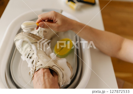 Close-up top view of male hands scrubbing muddy white sneakers with small brush and soap in white basin at home. Concept of shoe care, maintenance and cleanliness. Close-up top view of male hands scrubbing muddy white sneakers with small brush and soap in white basin at home. Concept of shoe care, maintenance and cleanliness. 127872533