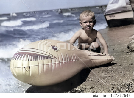 Vintage portrait of a little boy on the seashore. Boy near a big inflatable fish. Retro photo from 1968. Vintage portrait of a little boy on the seashore. Boy near a big inflatable fish. Retro photo from 1968. 127872648