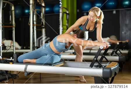 Girl trainer helps young woman to perform exercises in sitting position on pilates reformer Girl trainer helps young woman to perform exercises in sitting position on pilates reformer 127879514