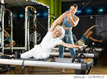 Girl woman trainer helps senior woman to perform exercises in sitting position on pilates reformer Girl woman trainer helps senior woman to perform exercises in sitting position on pilates reformer 127879517