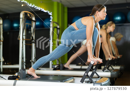 Young woman standing on pilates reformer, perform exercises Young woman standing on pilates reformer, perform exercises 127879530