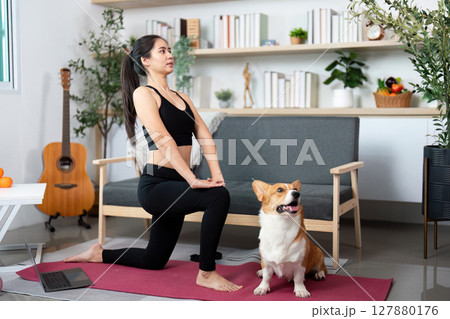 Yoga Session with Pet. Young woman in a yoga pose with her dog in a cozy home setting. 127880176