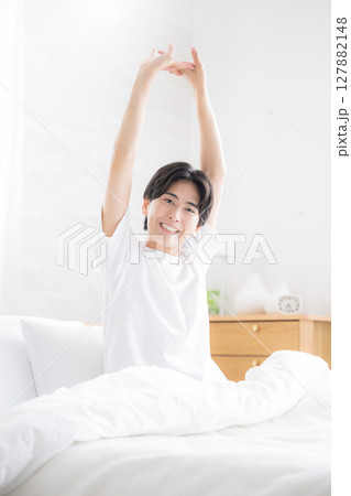 朝日を浴びるベッドで寝起きの若い男性　腕を伸ばしストレッチをする 127882148