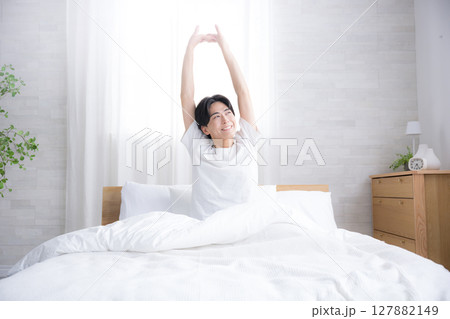 朝日を浴びるベッドで寝起きの若い男性　腕を伸ばしストレッチをする 127882149